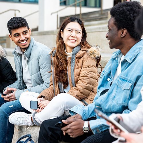 Eine Gruppe von jungen People of Colour sitzt auf einer Treppe. Zwei Frauen und ein Mann schauen dabei einen anderen Mann an und scheinen  zuzuhören. Sie lächeln dabei.
