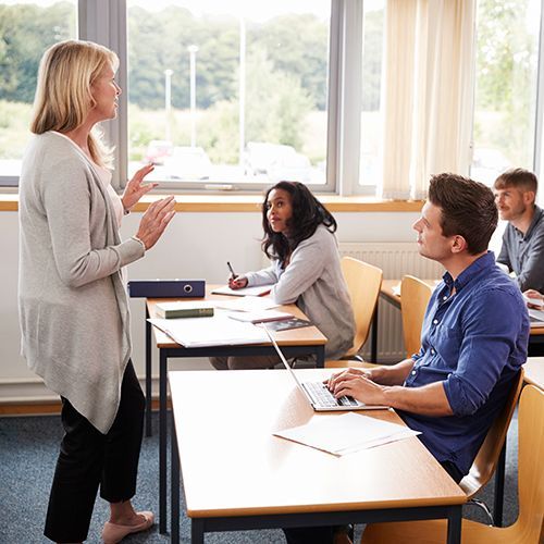 Eine Frau unterrichtet eine Gruppe von unterschiedlichen Menschen, die an Tischena lleine oder zu zweit sitzen. Zum Teil haben sie Laptops und Tablet-Computer vor sich.