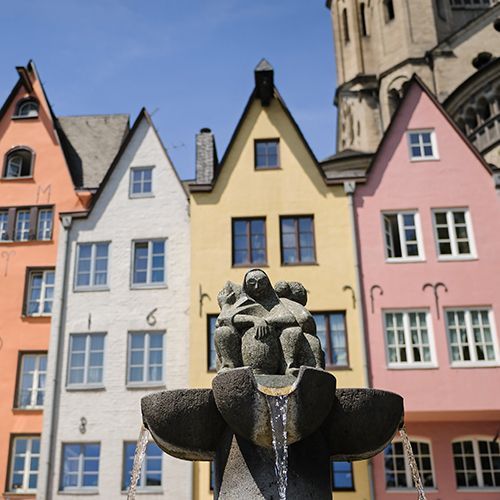 Fünf bunte Häuser in der Altstadt am Fischmarkt in Köln. Das linke Haus ist hellgrün, das daneben ist orange, dann folgt weiß, gelb und rosa. Im Vordergrund ist noch ein Brunnen zu sehen.