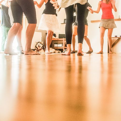 Junge Frauen und M&auml;nner stehen kreisf&ouml;rmig in einem gro&szlig;en Raum, halten sich an den H&auml;nden und tanzen miteinander.