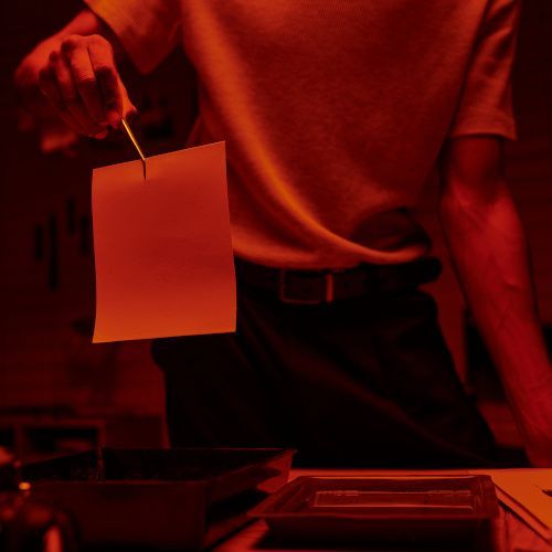 In einer Dunkelkammer die rot erleuchtet ist wird ein Fotoabzug mit einer Pinzette hochgehalten