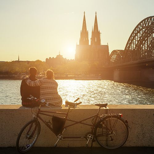 Ein P&auml;rchen sitzt auf einer Mauer am rechten Rheinufer in K&ouml;ln und schaut auf den Dom bei Sonnenuntergang. Die Hohenzollernbr&uuml;cke ist rechts zu sehen. Ein Tandem steht noch vor der Mauer.