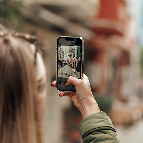 Eine Frau hält ein Smartphone um eine kleine Gasse zu fotografieren. Die Frau ist im Anschnitt an der linken Bildseite von hinten zu sehen.