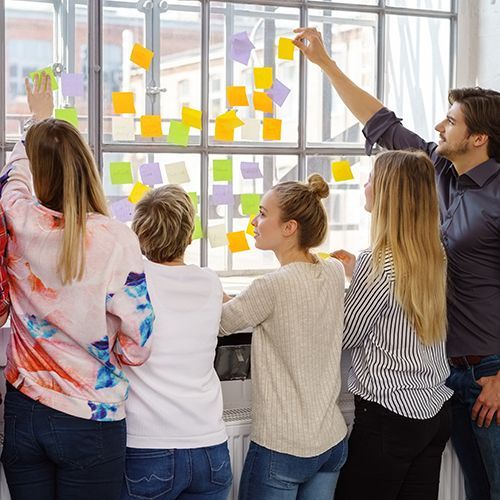 An einem gro&szlig;en Sprossenfenster stehen mehrere junge Menschen mit dem R&uuml;cken zur Kamera und kleben gelbe und gr&uuml;ne Post-its auf das Fenster.
