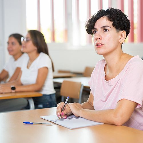 Eine Kursteilnehmerin sitzt an einem Tisch und notiert etwas mit Kugelschreiber in ein Heft. Im Hintergrund sind weitere Teilnehmer an Tischen zu sehen.