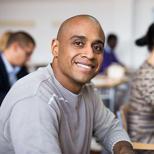 Ein Man of Colour schaut l&auml;chelnd in die Kamera. Im Hintergrund sind weitere Personen zu sehen die an Tischen sitzen und Stifte und Papier vor sich liegen haben.