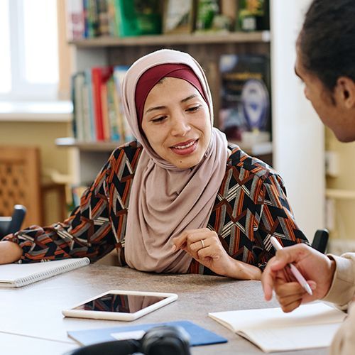 An einem Tisch sitzen drei People of Colour. Eine Frau mit Kopftuch beugt sich zu einem Mann und schaut auf dessen Unterlagen und er erkl&auml;rt ihr etwas. Auf dem Tisch liegen Bl&ouml;cke und ein Tablet