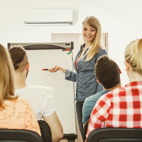 Eine weibliche Lehrkraft steht vor Teilnehmenden an einem Flipchart auf das sie mit einem roten Stift zeigt. Sie schaut die Teilnehmenden an.