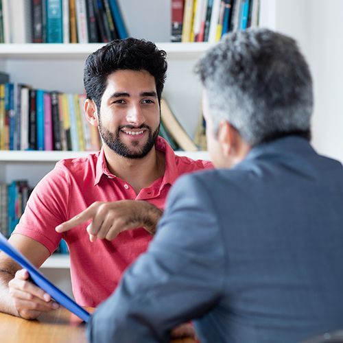 Ein Mann mit Bart zeigt auf einen Hefter einem anderen Mann der von hinten zu sehen ist. Im Hintergrund ist ein helles B&uuml;cherregal.