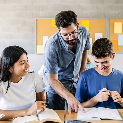 Zwei Teilnehmende sitzen an einem Tisch und haben B&uuml;cher vor sich aufgeschlagen. Ein Dozent beugt sich zwischen sie und zeigt einem Teilnehmer etwas im buch. Im Hintergrund sind Memo-Boards zu sehen.
