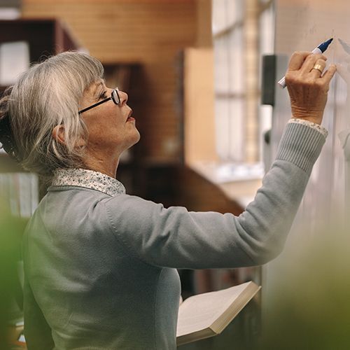 Eine Lehrkraft die an eine wei&szlig;e Tafel etwas schreibt. Die Lehrkraft tr&auml;gt zusammengesteckte graue Haare und eine Brille.