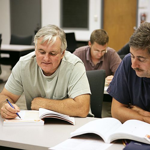 M&auml;nnliche Teilnehmende sitzen in einem Seminarraum zu zweit an Tischen . im Fokus sind zwei M&auml;nner zu sehen, die in ihr Buch schauen, das vor ihnen liegt. Ein Mann zeigt mit einem Stift ins Buch.