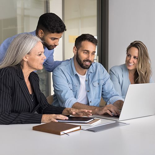 Drei Personen sitzen vor einem Laptop auf einem gro&szlig;en wei&szlig;en Tisch, eine weitere beugt sich dar&uuml;ber.