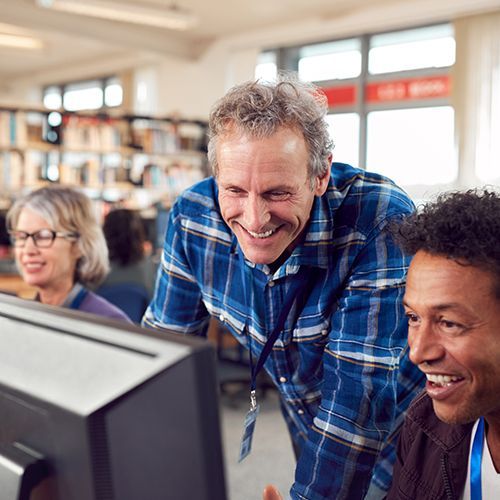 Ein Dozent mit einer Gruppe von Teilnehmenden eines Computer-Kurses. Der Dozent beugt sich &uuml;ber den Bildschirm eines Teilnehmers. Im Hintergrund sind unscharf B&uuml;cherregele zu sehen.