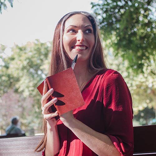 Eine &auml;ltere Frau mit langen braunen Haaren, zwei grauen Str&auml;hnen und einem roten Kleid steht drau&szlig;en und h&auml;lt ein Notizbuch in der Hand. Auch einen Stift h&auml;lt sie fest und sie schaut freudig als h&auml;tte sie etwas aufregendes vor.