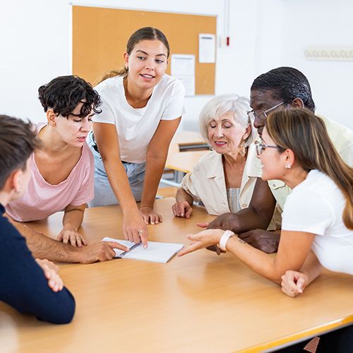 Eine Gruppe von unterschiedlichen Personen beugen sich &uuml;ber einen Zettel der in der Mitte auf einem Tisch liegt und diskutieren.