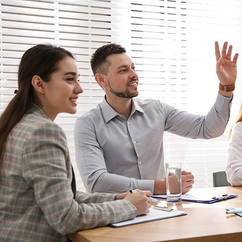 In einem hellen Raum stitzen an einem Holztisch mit Klemmbrettern drei Personen. Zwischen zwei Frauen sitzt ein Mann der die Hand hebt. Im Hintergrund sind die wei&szlig;en Jalousien eines gro&szlig;en Fensters zu sehen.