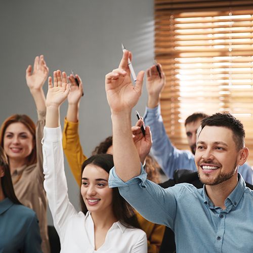 Mehrere Personen sitzen hintereinander in einem abgedunkelten Raum und heben die H&auml;nde zum Melden.