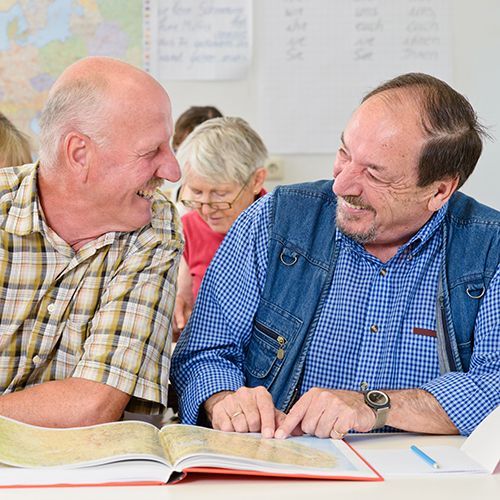 Zwei &auml;ltere M&auml;nner sitzen an einem Seminartisch zusammen und haben vor sich ein Buch aufgeschlagen. Sie schauen sich lachend an. Der linke Mann hat ein helles kariertes Hemd an und der rechte Mann ein blaukariertes. Im Hintergrund sind weitere Teilnehmer zu sehen.