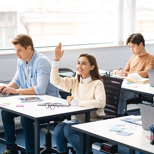 In einem hellen Seminarraum sitzen immer zwei Personan an Tischen die hintereinander stehen. Auf den Tischen sind Unterlagen, Tablets und Laptops. Eine junge Frau hebt l&auml;chelnd ihre Hand um sich zu melden.