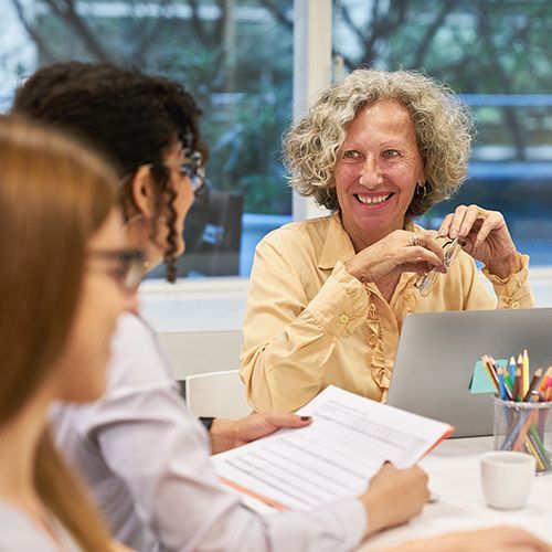 Drei Frauen sitzen an einem Tisch mit Laptop, Tablet und ein Glas mit Stiften. Zwei j&uuml;ngere Frauen sind seitlich angeschnitten, eine &auml;ltere Frau schaut diese lachend an und h&auml;lt ihre Brille in der Hand.