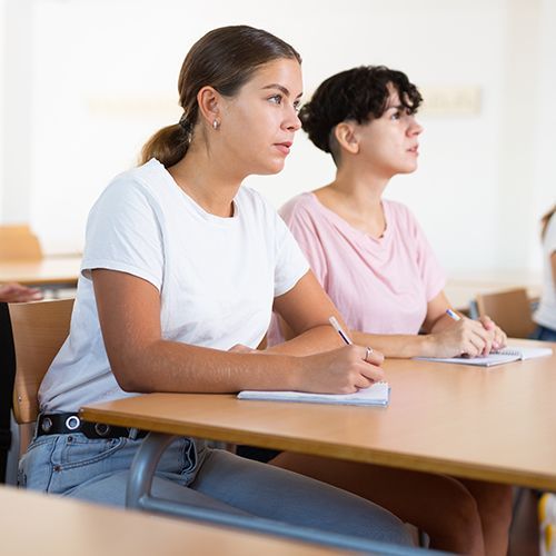 Mehrere Teilnehmer sitzen an Tischen und h&ouml;ren einer Person au&szlig;erhalb des Bildes zu und schauen dabei rechts aus dem Bild. Im Fokus ist eine junge Frau, die einen Stift in der Hand h&auml;lt und ein Heft vor sich liegen hat.