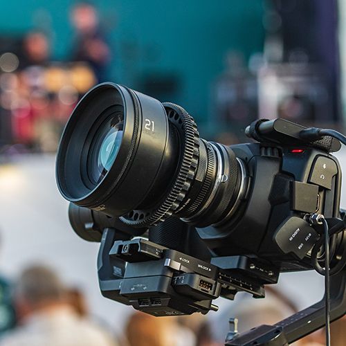 Eine profesionelle Kamera die  im rechten Bildrand positioniert ist und mit der Linse in die linke obere Rechte ausgerichtet ist. Im Hintergrund sind sehr unscharf Personen und eine B&uuml;hne zu sehen.