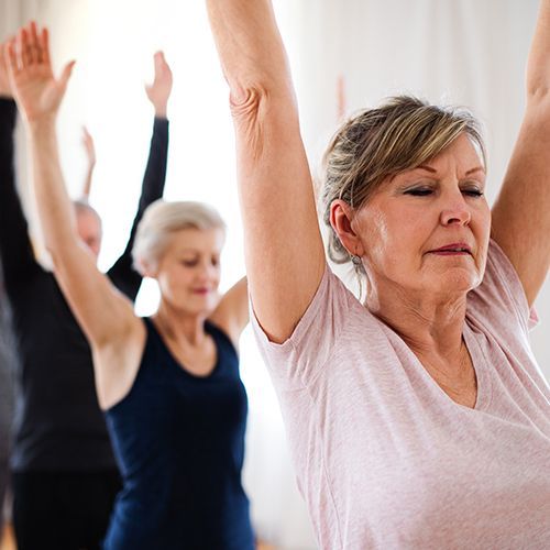 Drei Frauen und ein Mann um die 60 Jahre alt betreiben Sport. Sie stehen hintereinander in einer Reihe und heben die Arme nach oben.