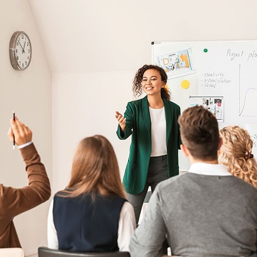 In einem Seminarraum sitzen Teilnehmende, eine Person meldet sich. Eine weibliche Lehrkraft steht an einem Whiteboard, das an der Wand h&auml;ngt.