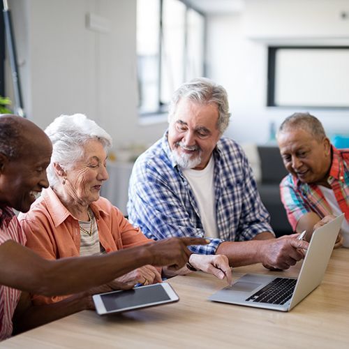Eine Gruppe Senioren sitzt gemeinsam an einem Tisch mit einem Lapt und einem Tablet. Die &auml;ltere Dame unter ihnen scheint sich von den &auml;lteren Herren den Umgang mit der Technik erkl&auml;ren zu lassen.