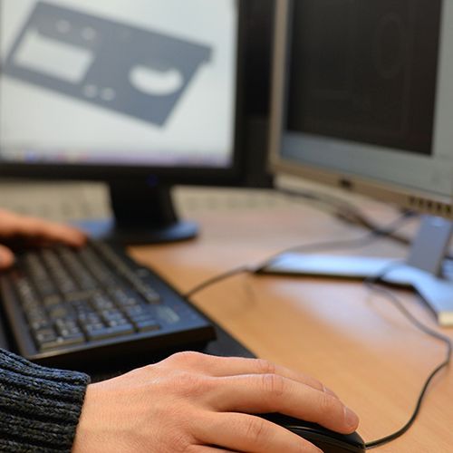 Zwei H&auml;nde sind zu sehen, die eine bedient eine PC-Maus, die andere liegt auf einer Tastatur. Im Hintergrund sind zwei Bildschirme, auf dem linken ist verschwommen eine Grafik abgebildet.