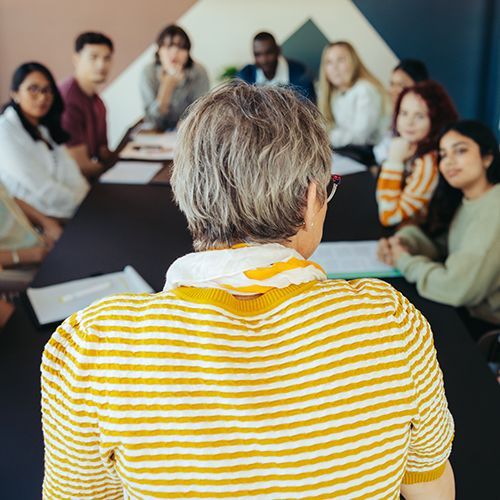 Teilnehmende sitzen an einem Tisch und schauen auf eine Dozentin, die mit dem R&uuml;cken zur Kamera steht.Sie tr&auml;gt ein gelb-wei&szlig; gestreiftes Shirt.