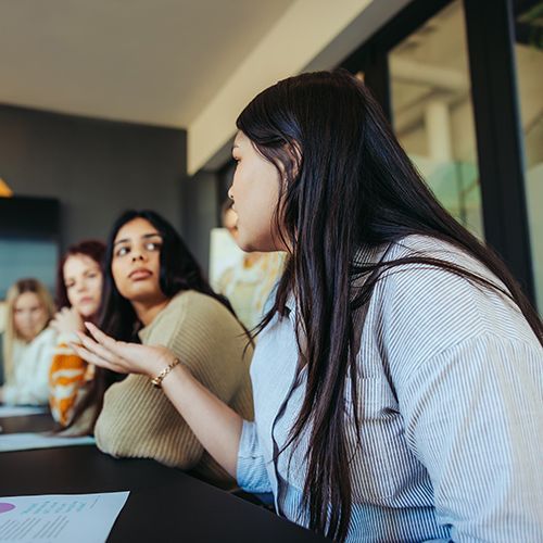 In einem Besprechungsraum sitzen mehrere Personen an einem langen dunklen Tisch. Sie sind von schr&auml;g unten aufgenommen. 
Im Fokus ist eine junge Frau mir langen schwarzen Haaren und einer gestreiften Bluse.  die zu den anderen hinhiseht, deswegen sieht man ihr Gesicht nicht richtig.