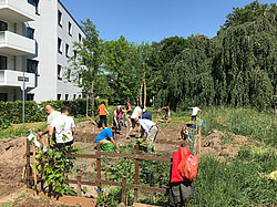 Erwachsene und Kinder pflegen gemeinsam eine Nutzfläche vor einem Mehrfamilienhaus
