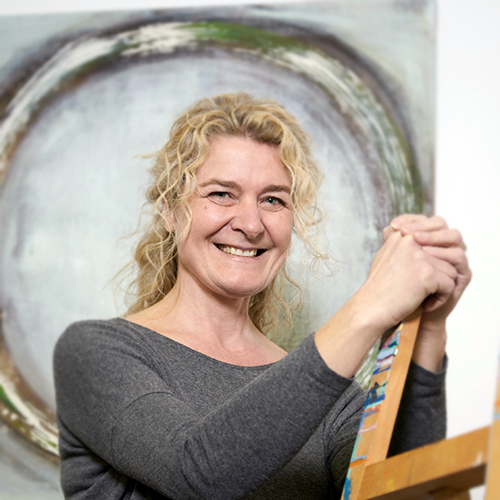 Portrait einer Dozentin mit blonden Locken, die sich an eine Staffeleil stützt.