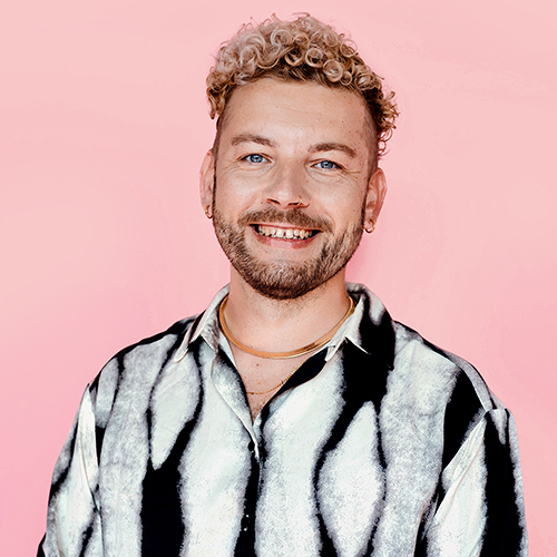 Ein Dozent mit blonden Locken steht vor einem rosafarbenen Hintergrund und lächelt in die Kamera